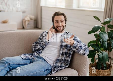 Felice attraente ragazzo caucasico millenario con stubble parla al telefono in soggiorno interno Foto Stock