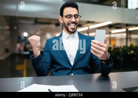 Felice attraente giovane uomo arabo con barba in occhiali, vestito a tavola guarda smartphone, fa gesto di vittoria Foto Stock