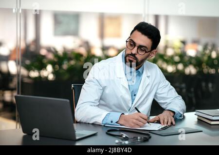 Serio attraente medico di tipo arabo millennial con la barba in occhiali, cappotto bianco guarda il laptop, fa note Foto Stock