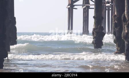 Sotto il molo in legno Crystal su pali, spiaggia oceano onde d'acqua, California USA. Vacanze estive sulla spiaggia di Mission, sulla riva di San Diego. Sotto il lungomare sulla costa del mare. Cinematografia senza giunture. Foto Stock