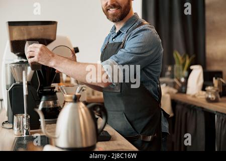 Il primo piano del barista versa i chicchi di caffè nel serbatoio della macchina da caffè per macinarli alla caffetteria Foto Stock