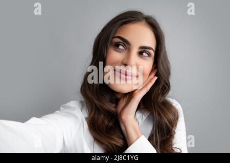 Ragazza piacevole facendo selfie in studio e ridendo. Giovane donna dall'aspetto bello con capelli ondulati marroni che scattano foto di se stessa su sfondo grigio. Foto Stock