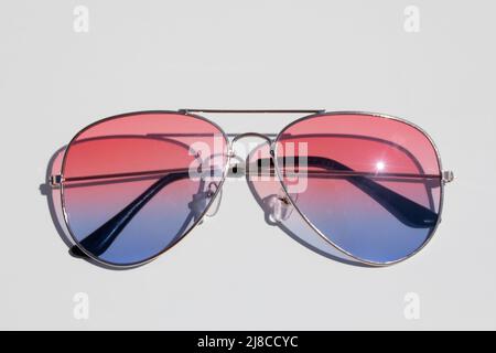 Gli occhiali da sole con lenti rosa e blu sono piatti su una superficie bianca. Illuminato da luce solare intensa. Concetto di vacanza e benessere. Foto Stock