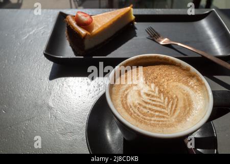 Vista dall'alto e ravvicinata di una tazza nera di cappuccino in ceramica con belle e delicate opere d'arte del latte con motivo a foglia e sfondo di torta sfocata. Foto Stock