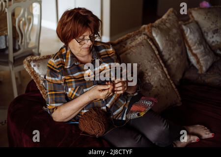 Donna con capelli rossi maturi in occhiali seduti sul divano in soggiorno e maglieria con aghi e palla di filo. Pensionato casa tempo libero e fatto a mano Foto Stock