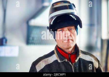Il saldatore maschio in abiti da lavoro con maschera sulla testa guarda direttamente nella macchina fotografica. Ritratto autentico di lavoratore in sala di produzione. Persone reali. Foto Stock
