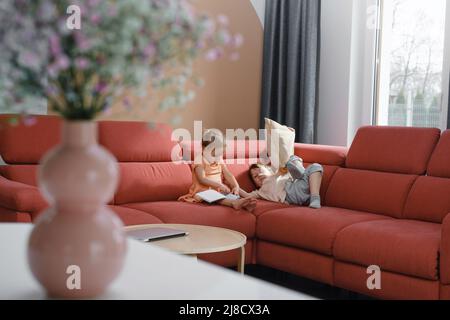 Fratello e sorella seduti sul divano. Fratelli e sorelle che comunicano a casa, giocano insieme. Famiglia Lifestyle. Vista anche se fiori in vaso. Moderno Foto Stock