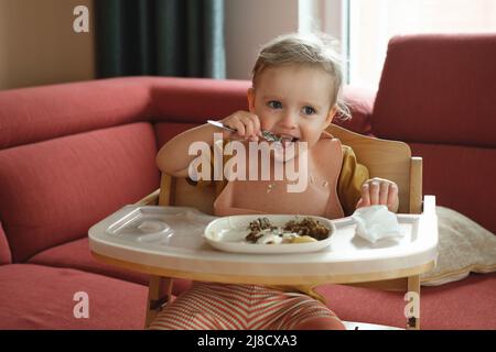 Bambina che mangia felicemente in cibo seggiolone da sola in camera. Bambini che mangiano. Buon bambino a pranzo. Bambina che si alimenta Foto Stock