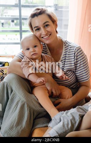 Madre con bambino nella camera dei bambini sopra la finestra. Ritratto di mamma e bambino a casa. Moderna famiglia. Bella mamma abbraccia neonato Foto Stock
