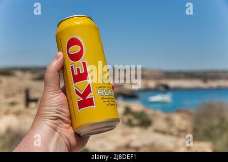 Ayia Napa, Cipro - 27 maggio 2021: La mano dell'uomo bianco tiene una lattina di birra KEO in primo piano con la costa estiva resort sullo sfondo. KEO è una birra cipriota, Foto Stock