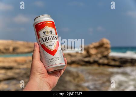 Ayia Napa, Cipro - 29 maggio 2021: La mano dell'uomo bianco tiene una lattina di birra argus lager vicino con Ayia Napa spiaggia resort estivo in background. Foto Stock