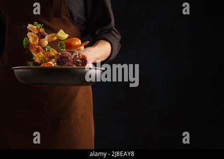 Uno chef professionista prepara frutti di mare - pezzi di pesce rosso, gamberi, polpo con verdure ed erbe in una padella su sfondo nero. Pubblicità, bann Foto Stock