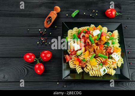 Penne pasta con verdure, piselli, peperoni dolci, pomodori, salsa, mozzarella su sfondo di legno nero. Pasta italiana con verdure e sp Foto Stock