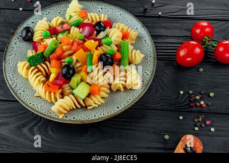 Penne pasta con verdure, piselli, peperoni dolci, pomodori, salsa, mozzarella su sfondo di legno nero. Pasta italiana con verdure e sp Foto Stock