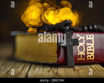 Un libro della Sacra Bibbia e un crocifisso su un rosario su un semplice tavolo di legno su sfondo giallo con luci scintillanti. Gesù Cristo, religione Foto Stock