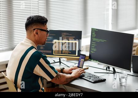 Vista laterale dello sviluppatore IT asiatico che digita sulla tastiera con il codice di programmazione sullo schermo del computer mentre lavora in ufficio Foto Stock