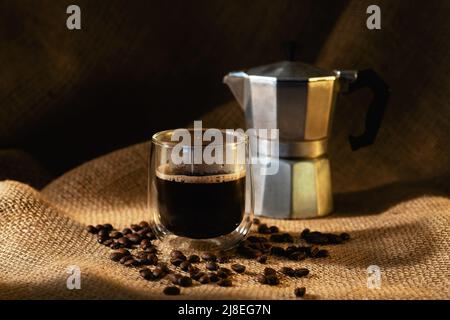 Una tazza di caffè in vetro trasparente e una macchina per il caffè geyser su sfondo scuro con chicchi di caffè. Foto orizzontale. Foto Stock