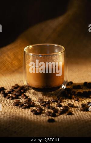 Una tazza di caffè in vetro trasparente con latte su sfondo scuro con chicchi di caffè. Foto Stock