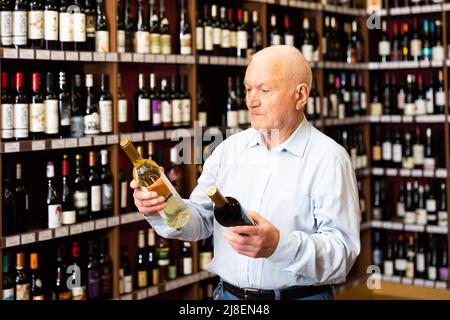 Un anziano serio sceglie tra vino rosso e bianco in un negozio di liquori Foto Stock