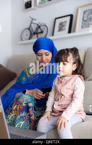 Piccolo bambino che guarda i cartoni animati sul computer portatile. Madre e figlia insieme. Famiglia musulmana single-parente a casa. Foto Stock