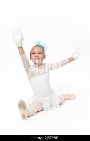 Ballerina caucasica girl in gonna di balletto bianco tutu, arco blu in capelli e balletto di mittens bianco che si allunga su pavimento bianco. Foto studio Foto Stock