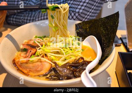 I chopsticks raccolono il ramen giapponese del tonkotsu con le alghe, il maiale (tonkotsu), il germoglio del fagiolo e gli scallioni in una ciotola. Foto Stock