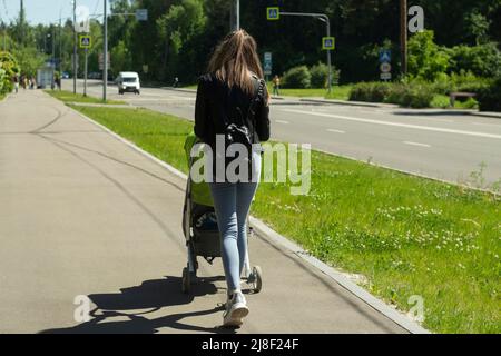 Una ragazza con un passeggino con un bambino. Una donna cammina con un bambino. La ragazza cammina lungo la strada con un passeggino. Foto Stock