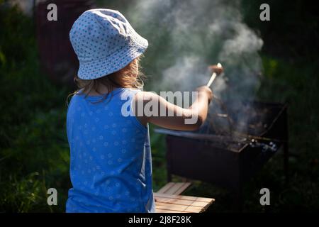 Bambini al picnic. La ragazza cuoce il pane sul bastone. Attività ricreative estive all'aperto. Bambino vicino al fuoco. Bambino a panama. La ragazza tiene il bastone di pane sopra il fuoco. Foto Stock