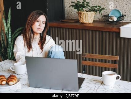Donna bruna sorridente adulta che fa colazione con un computer portatile aperto in cucina a casa Foto Stock