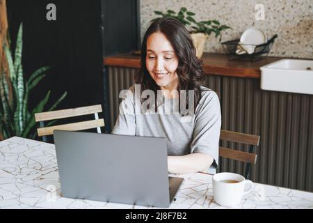 Donna bruna sorridente adulta che fa colazione con un computer portatile aperto in cucina a casa Foto Stock