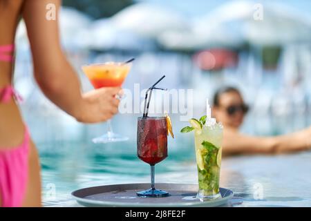 Due cocktail freschi e freschi su un vassoio rotondo vicino alla piscina. Vista crop della mano femminile che tiene un bicchiere con bevanda mentre si rilassa in piscina, con resort sfocato sullo sfondo. Concetto di rinfresco. Foto Stock