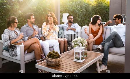 Gruppo di giovani allegri seduti insieme bevendo vino nel giardino di lusso - gli amici si riuniscono seduti su un divano nel retro della terrazza Foto Stock