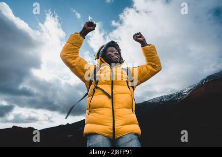 Un giovane escursionista di etnia africana in un gilet giallo e zaino si rallegra con le sue braccia sollevate - concetto di vittoria, di girovagosta e di successo. Foto Stock
