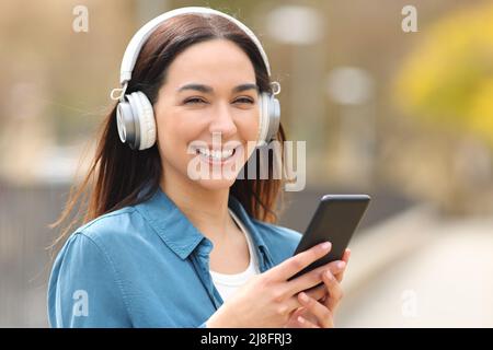 Una donna felice che indossa cuffie con smartphone ti guarda in un parco Foto Stock