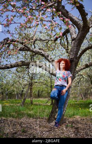 Curly donna contadina rossa nel suo giardino di mele in piena fioritura Foto Stock