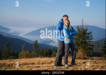 Giovane uomo che abbraccia la sua ragazza divertente da dietro mentre si trova in piedi nel prato di montagna in autunno giorno di sole. Paesaggio affascinante delle leggendarie montagne in nebbia dietro di loro. Foto Stock