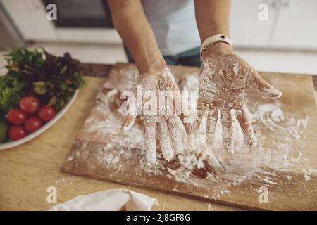 Le mani delle donne in farina sullo sfondo di un tavolo da cucina e di un piatto di verdure Foto Stock