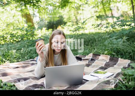 donna sorridente in occhiali sdraiati sulla coperta, tenendo la tazza di carta e utilizzando il computer portatile nel parco Foto Stock