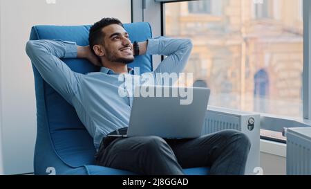Giovane professionista arabo che lavora su un portatile, sogna, crea una nuova idea. Sorridente dipendente indiano in professione creativa si siede a proprio agio Foto Stock