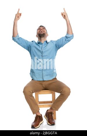 uomo entusiasta con pantaloni chino che guardano verso l'alto e puntano le dita nell'aria, essendo grato e ringraziando dio di fronte a sfondo bianco in studio Foto Stock