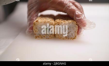 Il primo piano di un guanto taglia i rotoli, il processo di fabbricazione dei rotoli. Il processo di preparazione del sushi giapponese. La lama in mano taglia un primo piano del rullo su un legno Foto Stock