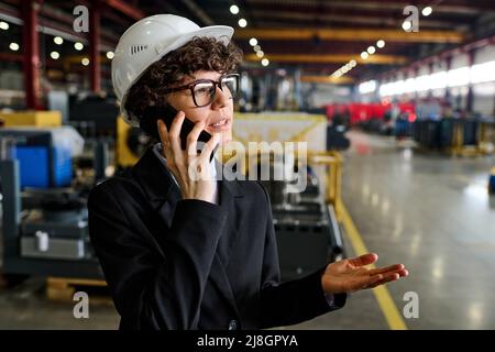 Giovane manager fiducioso della fabbrica moderna che parla al telefono cellulare mentre si consulta un collega o subordina in un'officina di grandi dimensioni Foto Stock