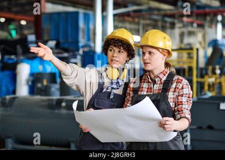 Giovane ingegnere fidato donna che punta a nuove attrezzature durante l'incontro con la collega con il progetto mentre spiega qualcosa Foto Stock