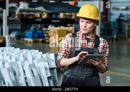 Giovane ingegnere femminile di fabbrica moderna con tablet che controlla la qualità delle nuove apparecchiature industriali e l'inserimento dei dati nel database Foto Stock