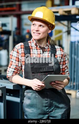 Giovane sorridente lavoratrice di fabbrica che lavora presso apparecchiature industriali e utilizza tablet mentre lavora con i dati in magazzino Foto Stock