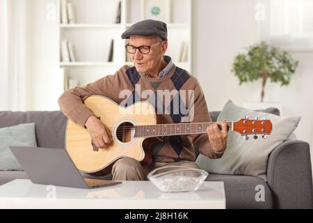 Uomo anziano che suona una chitarra acustica e si siede di fronte a un computer portatile a casa Foto Stock