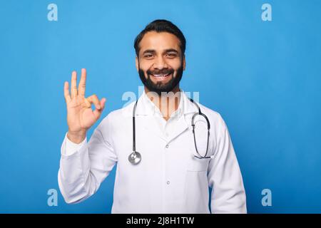 Amichevole medico indiano allegro indossare abito bianco e stetoscopio mostrando segno ok con la mano e le dita, guardando la fotocamera con un sorriso, uomo in uniforme medico isolato su blu Foto Stock