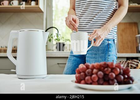 Tempo del tè, Donna versare acqua bollita dal bollitore elettrico in una tazza per preparare il tè, la prima colazione del mattino in cucina Foto Stock