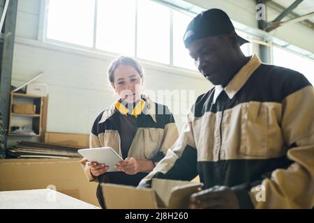 Giovane donna in uniforme utilizzando il tablet digitale mentre la sua collega confezionava la scatola al tavolo Foto Stock