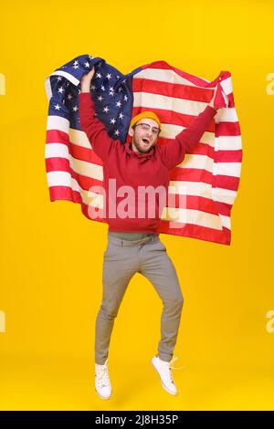 Giovane allegro con bandiera americana. Felice, divertente giovane uomo con cappuccio rosso e bandiera USA che saltano hight isolato su sfondo giallo. La libertà è nella tua vita. Foto Stock